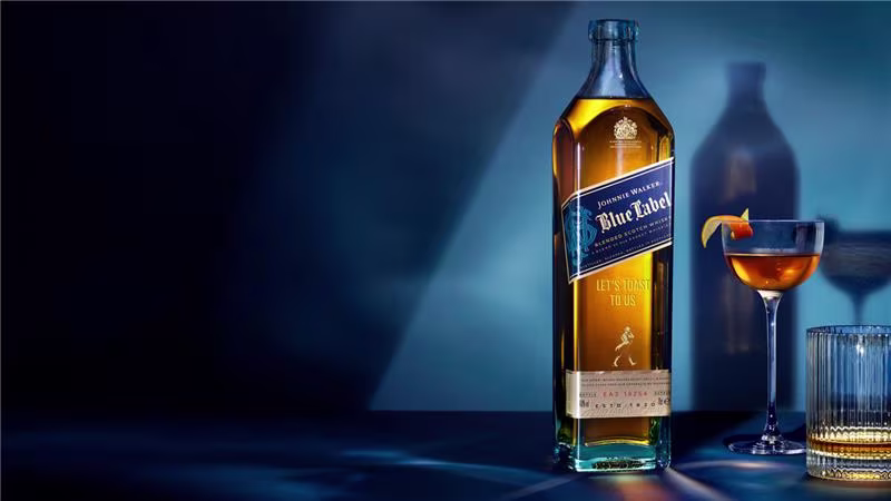 一瓶高高的尊尼獲加藍牌威士忌，旁邊放著一個飾有橙皮的雞尾酒杯和一杯水，以藍色背景和戲劇性的燈光為襯托。