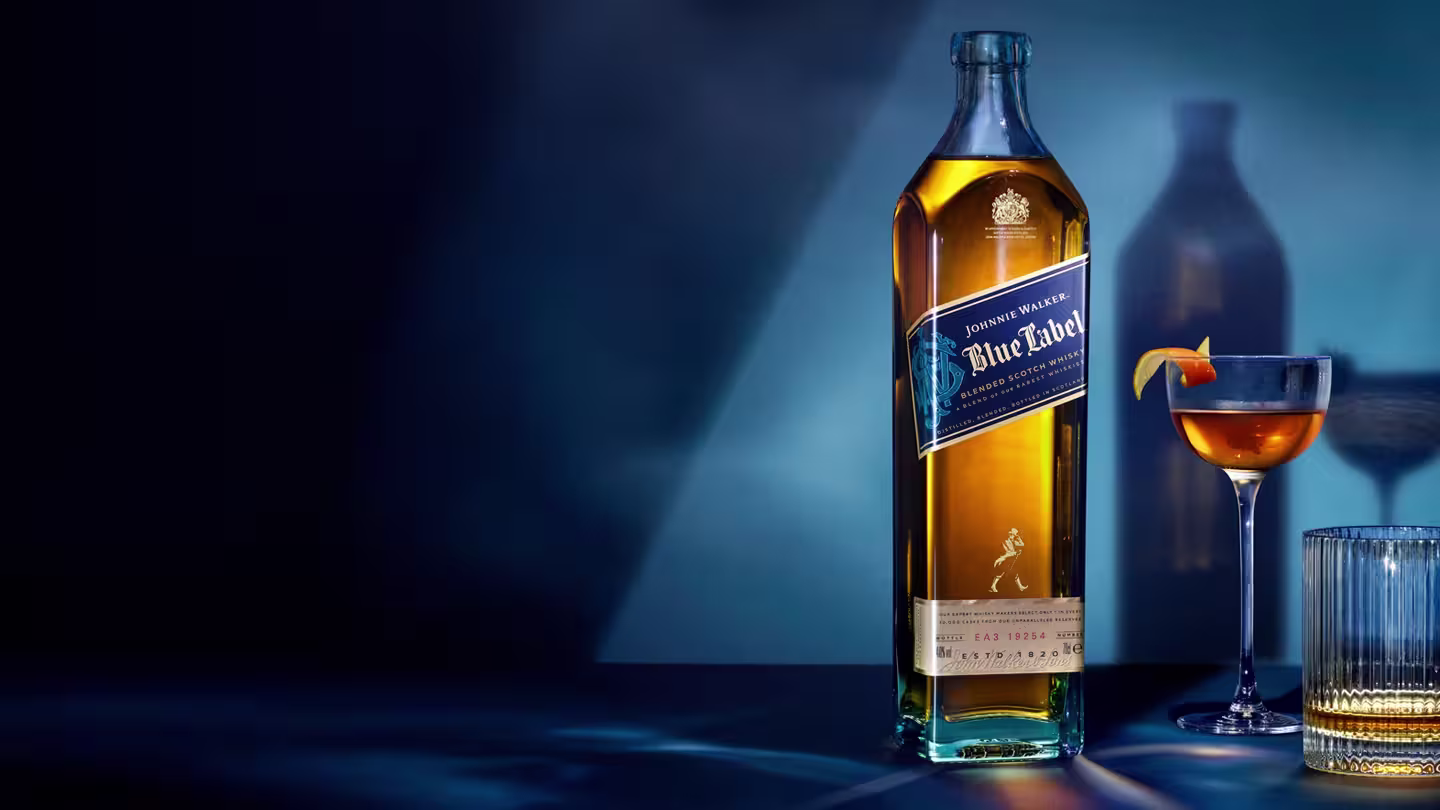 Eine Flasche Johnnie Walker Blue Label Whisky steht auf einem Tisch neben einem Cocktailglas, garniert mit einer Orangenschale, und einem Glasbecher, alles vor einem dunkelblauen Hintergrund mit dramatischer Beleuchtung.
