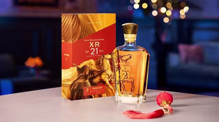 一瓶尊尼獲加 XR 21 年調和蘇格蘭威士忌，旁邊擺放著與之配套的金色盒子，前面掛著紅色裝飾流蘇，背景是模糊的節日彩燈。