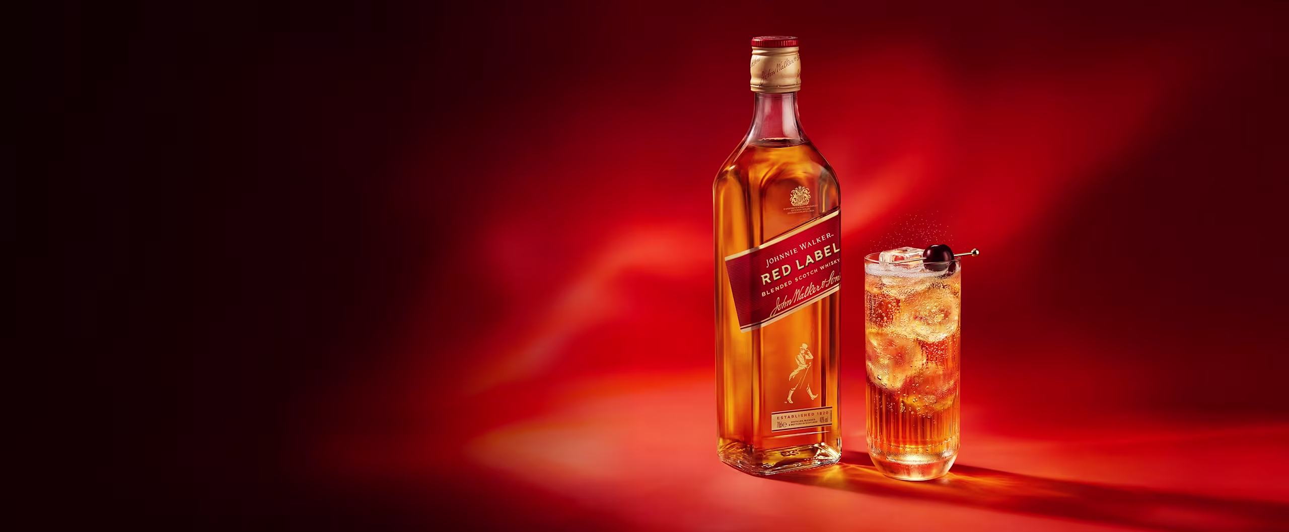 A bottle of Johnnie Walker Red Label whisky next to a glass of whisky with ice on a red background.