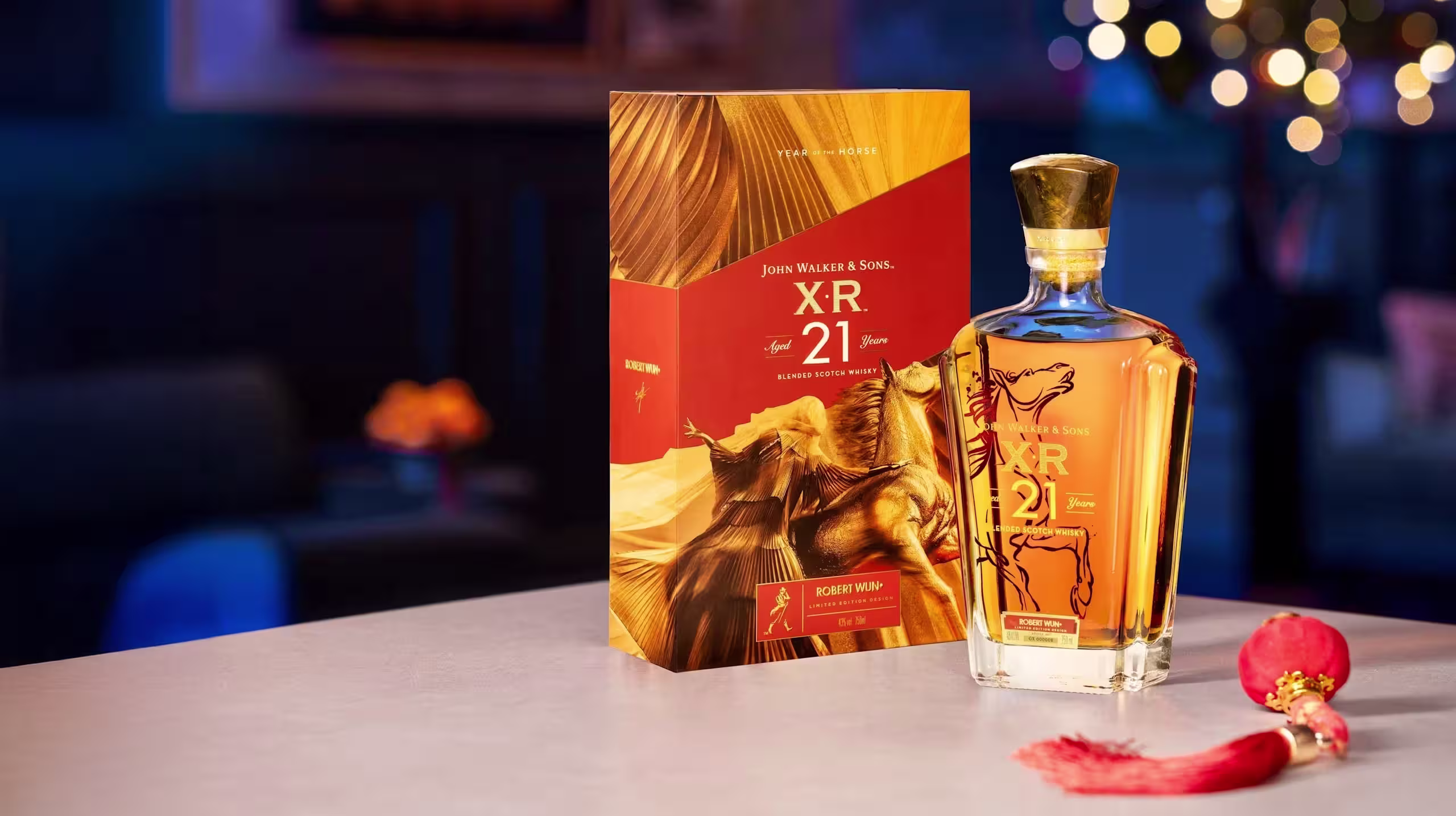 一瓶尊尼獲加 XR 21 年威士忌擺在桌上,旁邊是它的裝飾盒,附近有一個紅色流蘇裝飾品,背景是模糊的節日彩燈。