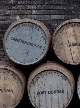 Stacked wooden barrels against a dark brick wall, featuring labels such as Cameronbridge and Port Dundas from John Walker & Sons.