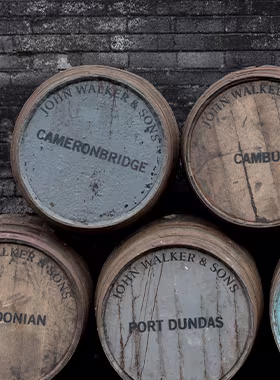 Gros plan de cinq barils en bois empilés, chacun étiqueté « John Walker & Fils ». Les noms visibles sur les barils sont Cameronbridge, Cambus, Bonian et Port Dundas. Les barils sont disposés contre un mur de briques sombres.