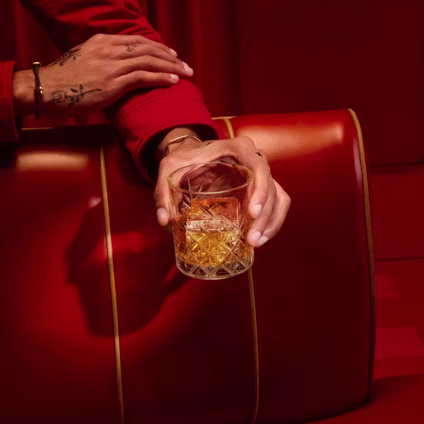 Una persona vestida de rojo, mostrando manos tatuadas, sostiene un vaso de bebida de color ámbar mientras descansa en un sofá de cuero rojo con ribetes amarillos.