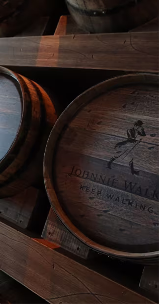 A close-up of several wooden whiskey barrels stacked in a dimly lit warehouse. The barrels are weathered and show signs of use, with visible grooves and markings. The logo of a well-known whiskey brand is burned into the side of one of the barrels.