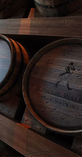A close-up of several wooden whiskey barrels stacked in a dimly lit warehouse. The barrels are weathered and show signs of use, with visible grooves and markings. The logo of a well-known whiskey brand is burned into the side of one of the barrels.