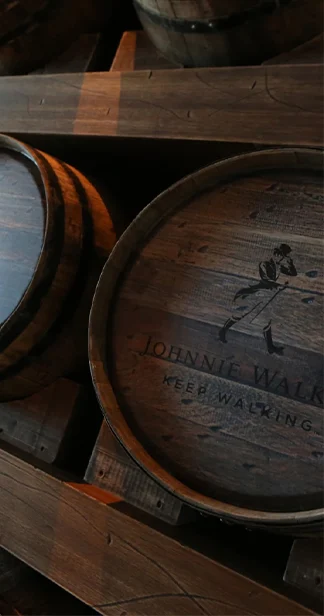 A close-up of several wooden whiskey barrels stacked in a dimly lit warehouse. The barrels are weathered and show signs of use, with visible grooves and markings. The logo of a well-known whiskey brand is burned into the side of one of the barrels.