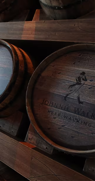 Дървени бъчви, подредени в тъмна обстановка, с текст JOHNNIE WALKER ПРОДЪЛЖАВАЙТЕ ДА ВЪРВИТЕ и емблематичното лого на ходещ човек върху един от капаците на бъчвите.
