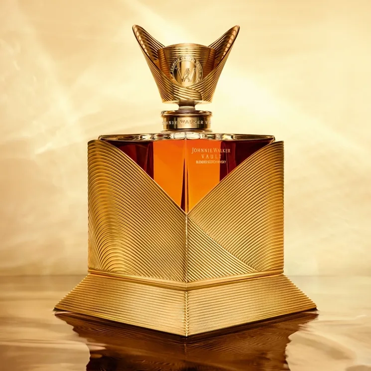 A luxurious gold bottle of Johnnie Walker whisky with a sculpted, geometric design sits against a shimmering golden background. The intricate bottle features bold, wavy lines and a decorative cap.