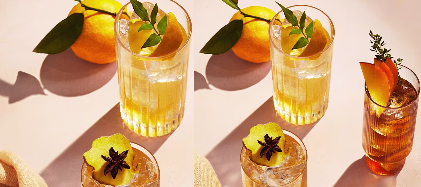 Two Johnnie Walker whisky cocktails in clear glasses on a table. The left drink is garnished with a star anise and apple slice, while the right has orange peel and thyme. A whole orange with green leaves is in the background, casting a shadow.