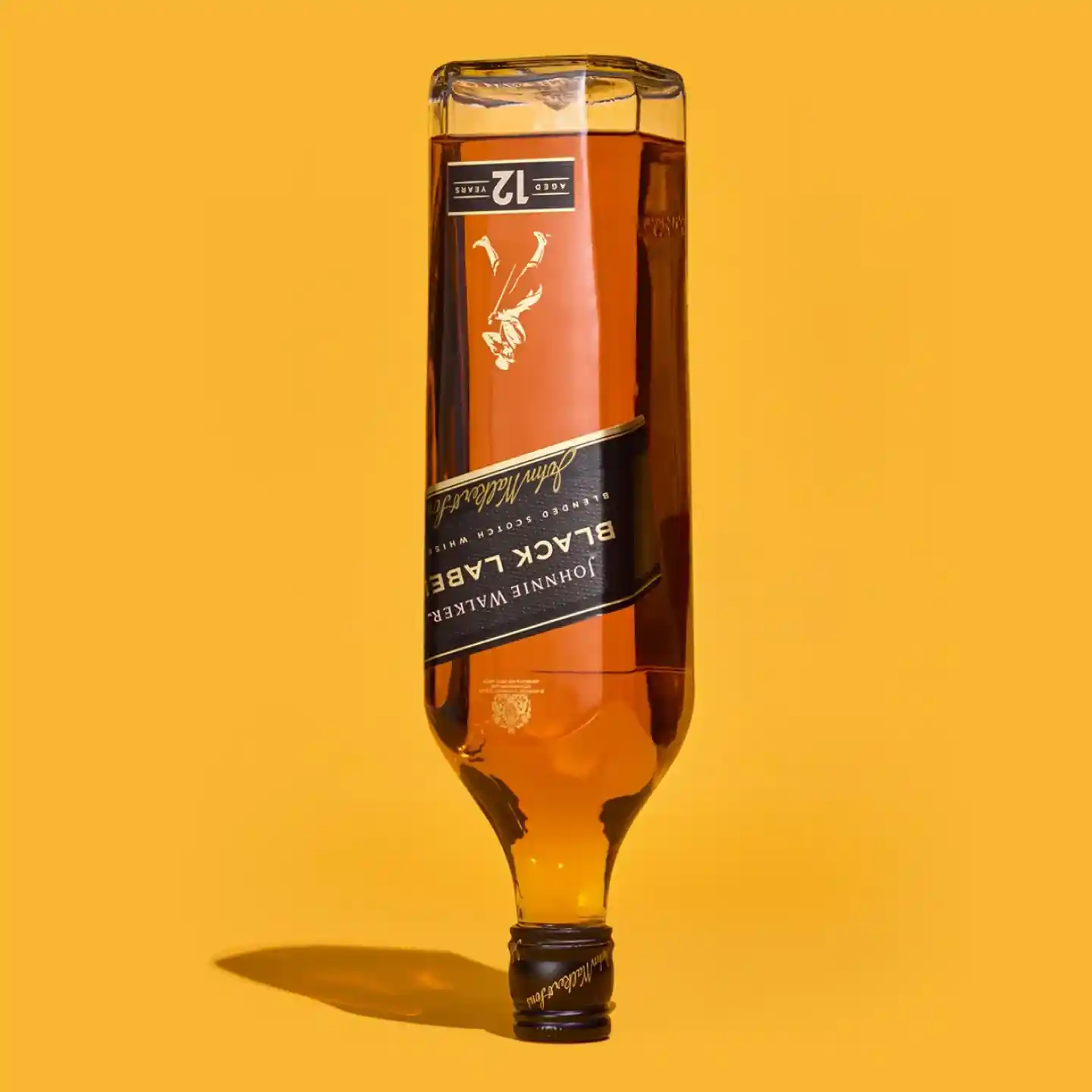 Převrácená láhev whisky Johnnie Walker Black Label stojí na jejím uzávěru na pevném oranžovém pozadí. Štítek a kapalina uvnitř jsou jasně viditelné.