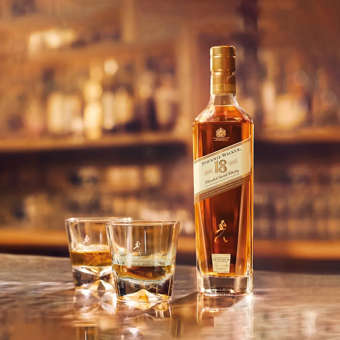 A bottle of Johnnie Walker 18-year-old blended Scotch whisky is on a bar counter, accompanied by two glasses filled with the whisky. The background features a blurred view of a warm, dimly lit bar setting.