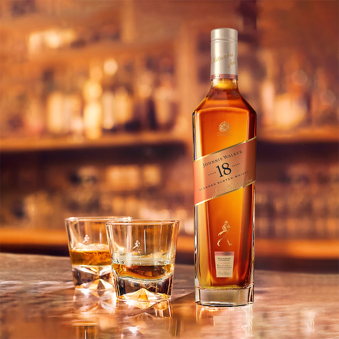 Ένα μπουκάλι 18 ετών blended σκωτσέζικο ουίσκι Johnnie Walker στέκεται σε μια μπάρα δίπλα σε δύο ποτήρια γεμάτα ουίσκι, με ένα ελαφρώς θολό φόντο.