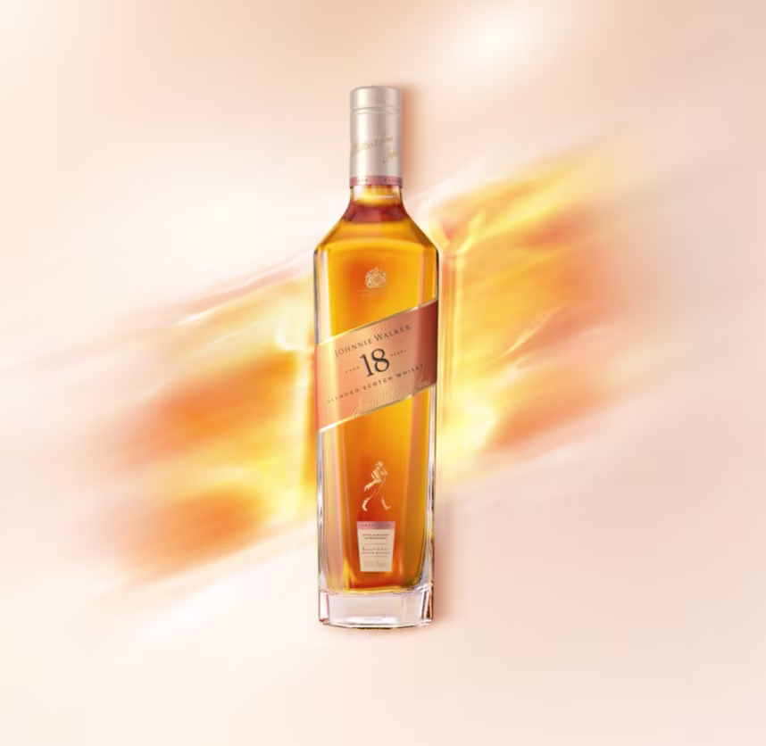 A bottle of Johnnie Walker 18-year-old blended Scotch whisky is on a bar counter, accompanied by two glasses filled with the whisky. The background features a blurred view of a warm, dimly lit bar setting.