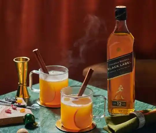 Dos jarras de cristal que contienen bebidas calientes y especiadas con ramas de canela, junto a una botella de whisky Johnnie Walker Black Label. Sobre una mesa verde hay un medidor, un mantel y una tabla con adornos. 