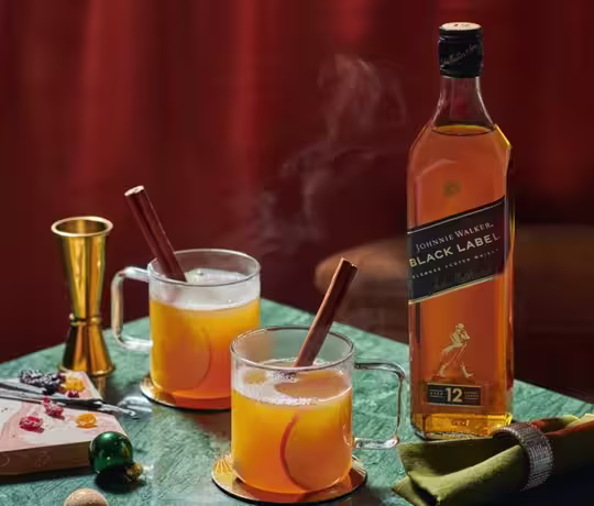 Two glass mugs with a warm beverage garnished with cinnamon sticks, placed on a green table. Beside them is a bottle of Johnnie Walker Black Label whisky. A jigger and some sweets are also on the table, with an orange and green cloth nearby.