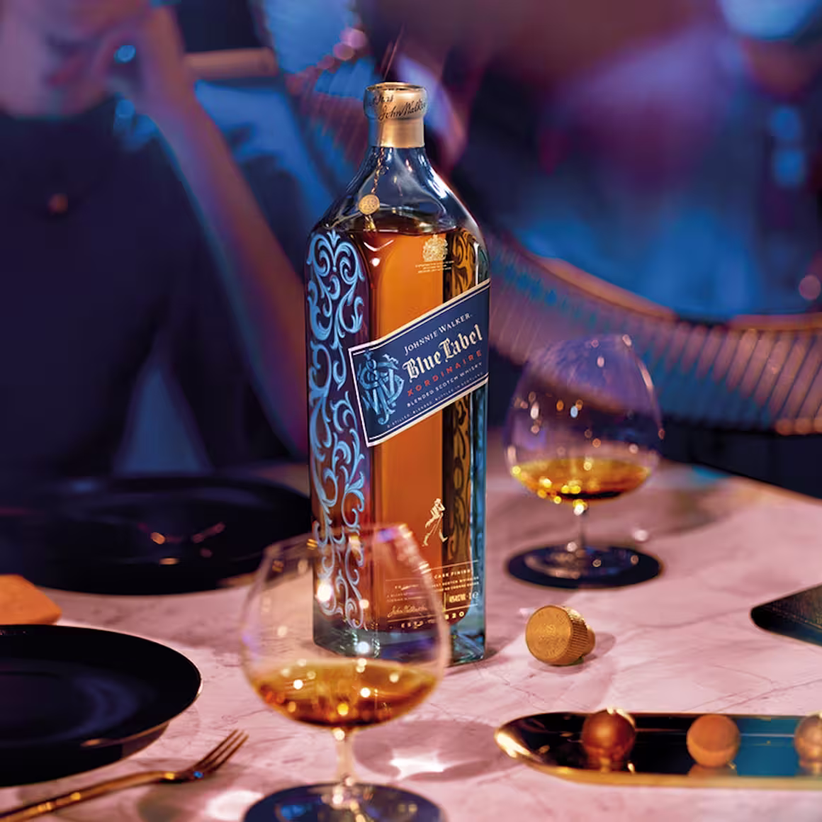 A bottle of Johnnie Walker Blue Label whisky stands on a marble table with two glasses of whisky, a cork stopper, and black plates, in a dimly lit, elegant setting.