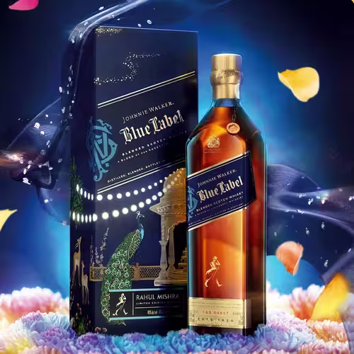 A bottle of Johnnie Walker Blue Label whisky stands beside an ornate, dark blue box with gold and peacock designs. Colorful flower petals float around on a deep, vibrant background.