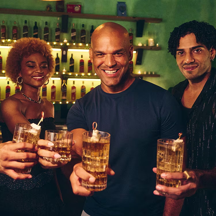 Tres personas sonriendo y brindando con cócteles en un bar cálidamente iluminado, con estanterías de botellas al fondo.