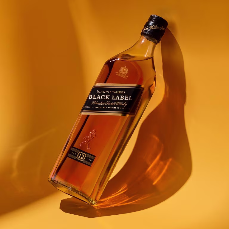  A bottle of Johnnie Walker Black Label 12 Year Old Blended Scotch Whiskey is tilted against a yellow background. The bottle casts a shadow, and the label is clearly visible with a black cap.