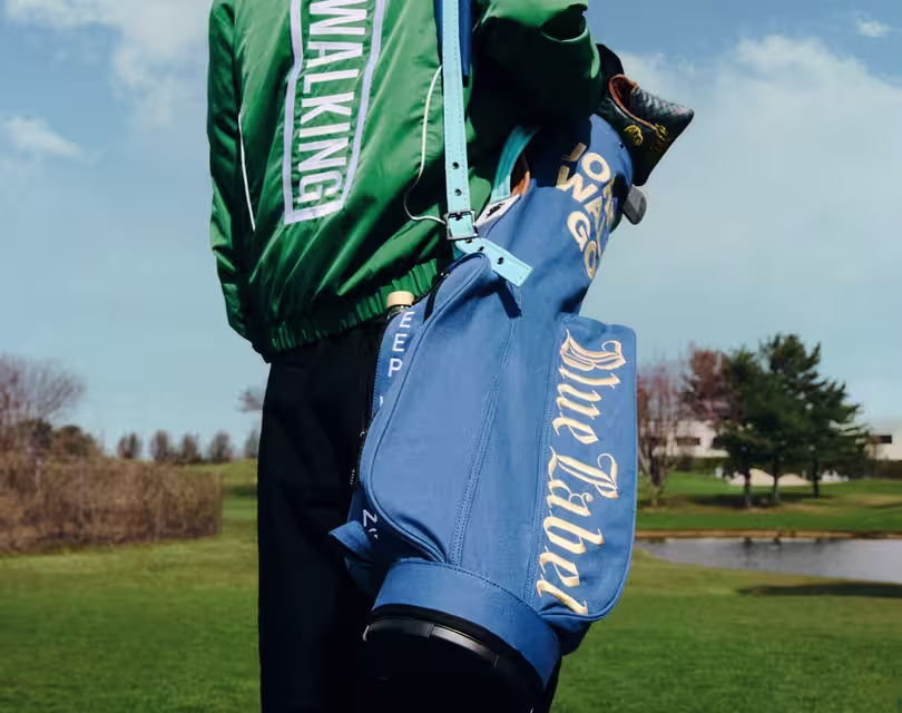 A person in a green jacket with WALKING written on the sleeve carries a blue golf bag over their shoulder. The golf bag has Blue Label written on it. They stand on a grassy field with trees and a pond in the background.