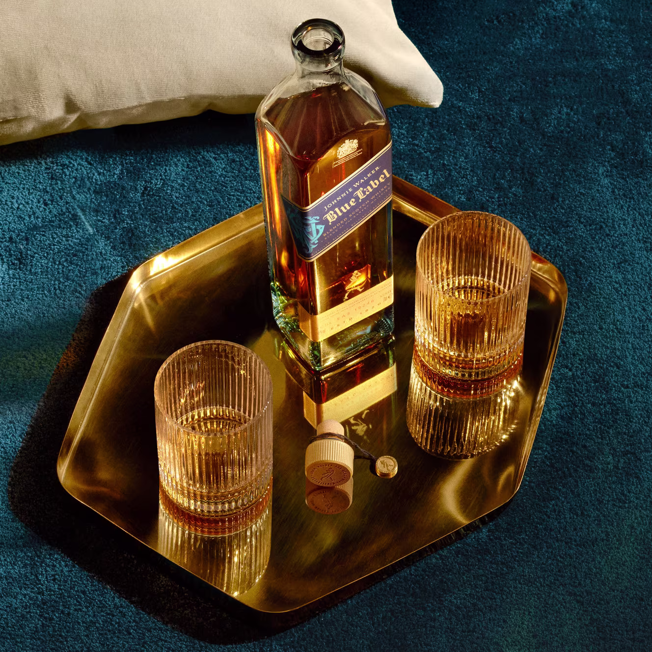 A bottle of Blue Label whiskey and two filled glasses sit on a hexagonal gold tray atop a blue textured surface. The bottles cap and a coin are next to the glasses, with a pillow partially visible in the top left corner.