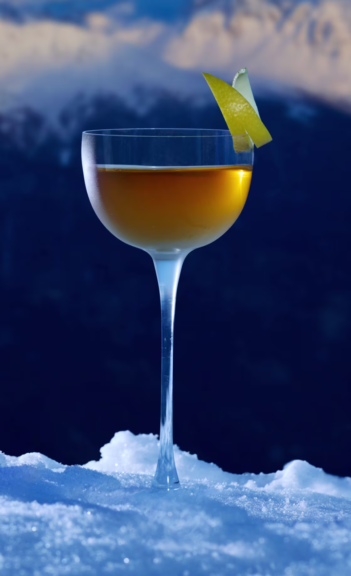A Ice Chalet Manhattan cocktail in a coupe glass with a lemon twist garnish is placed on snow. The background features blurred mountain peaks under a clear blue sky, highlighting the drink’s golden hue.