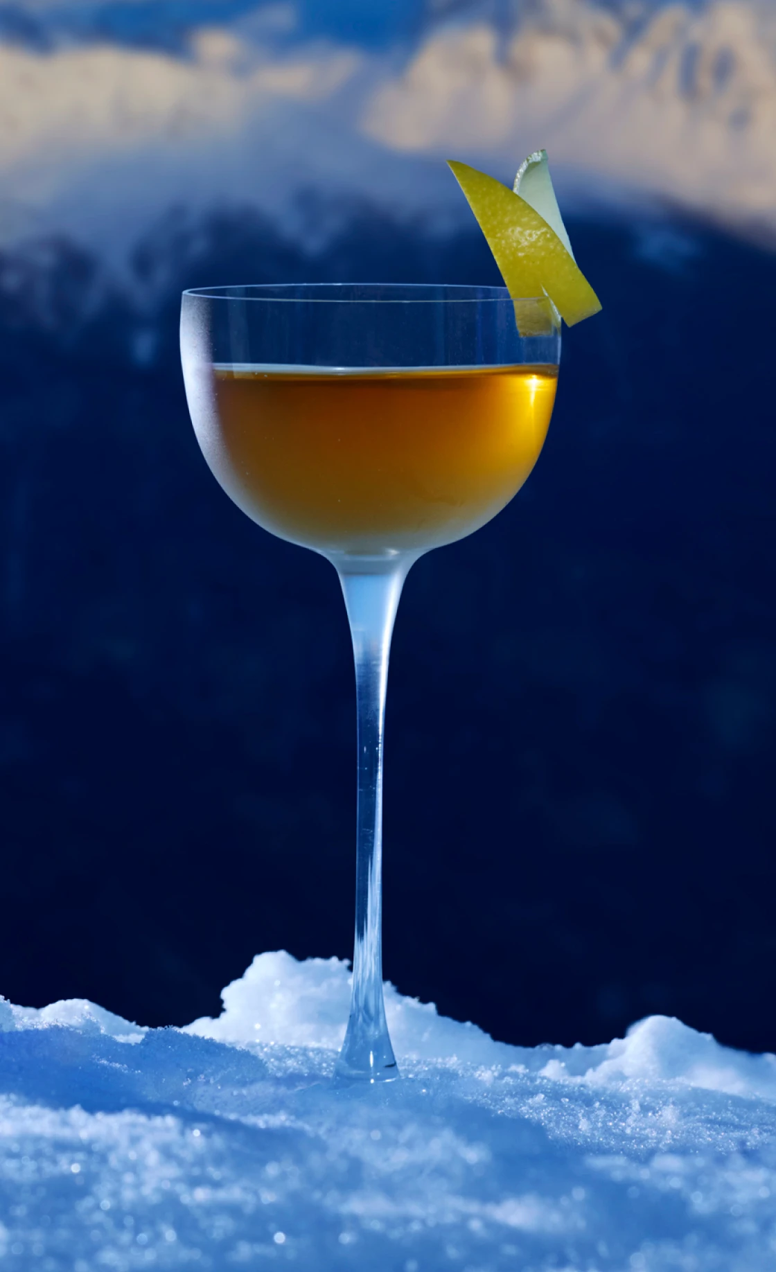 A Ice Chalet Manhattan cocktail in a coupe glass with a lemon twist garnish is placed on snow. The background features blurred mountain peaks under a clear blue sky, highlighting the drink’s golden hue.