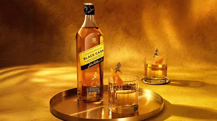 A bottle of Johnnie Walker Black Label whiskey stands on a golden tray next to two glasses of whiskey with orange peels, all set against a warm golden background.