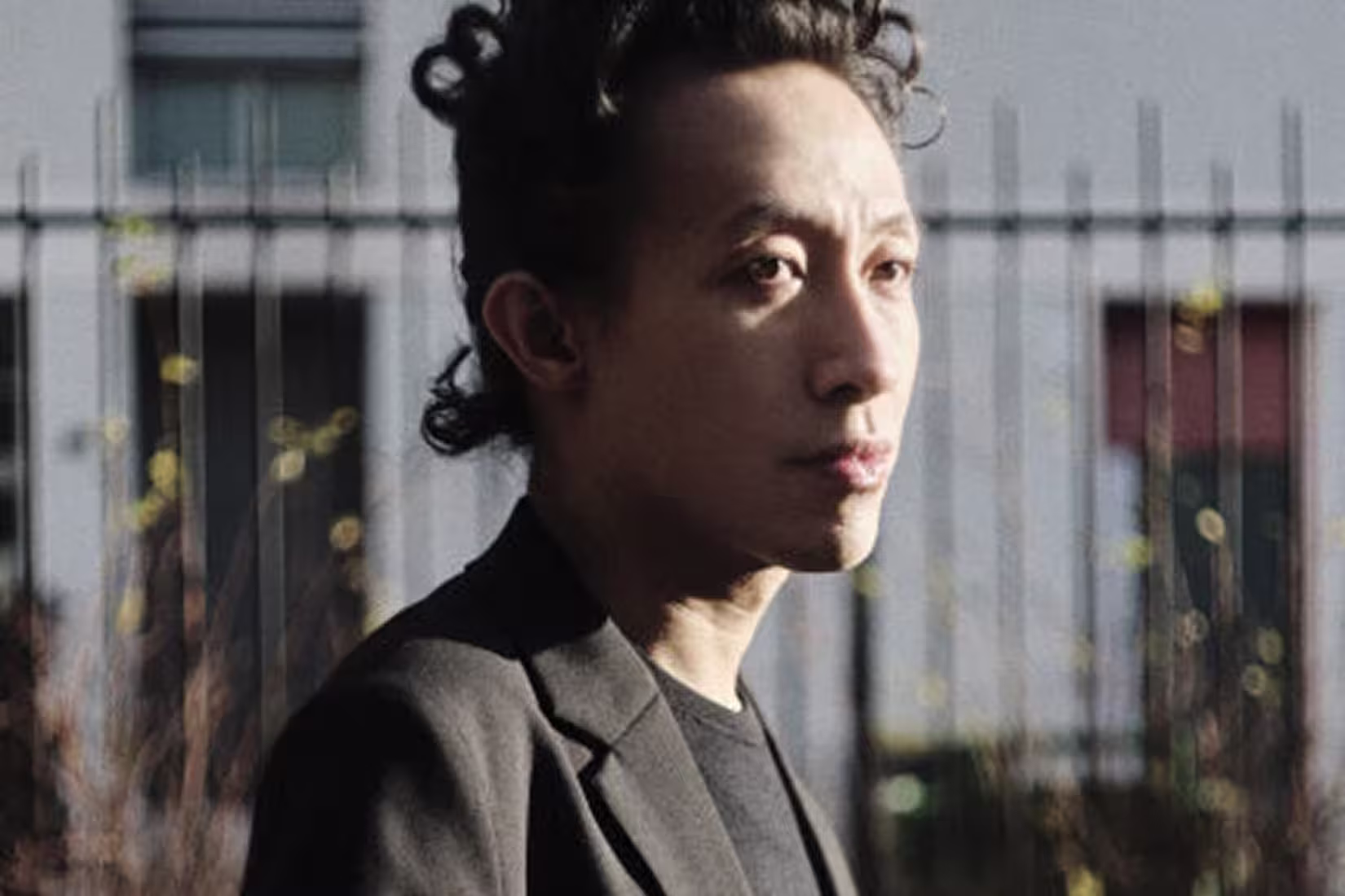 A person with curly dark hair wearing a black jacket stands outdoors in sunlight, looking to the side. A metal fence and blurred building are visible in the background.