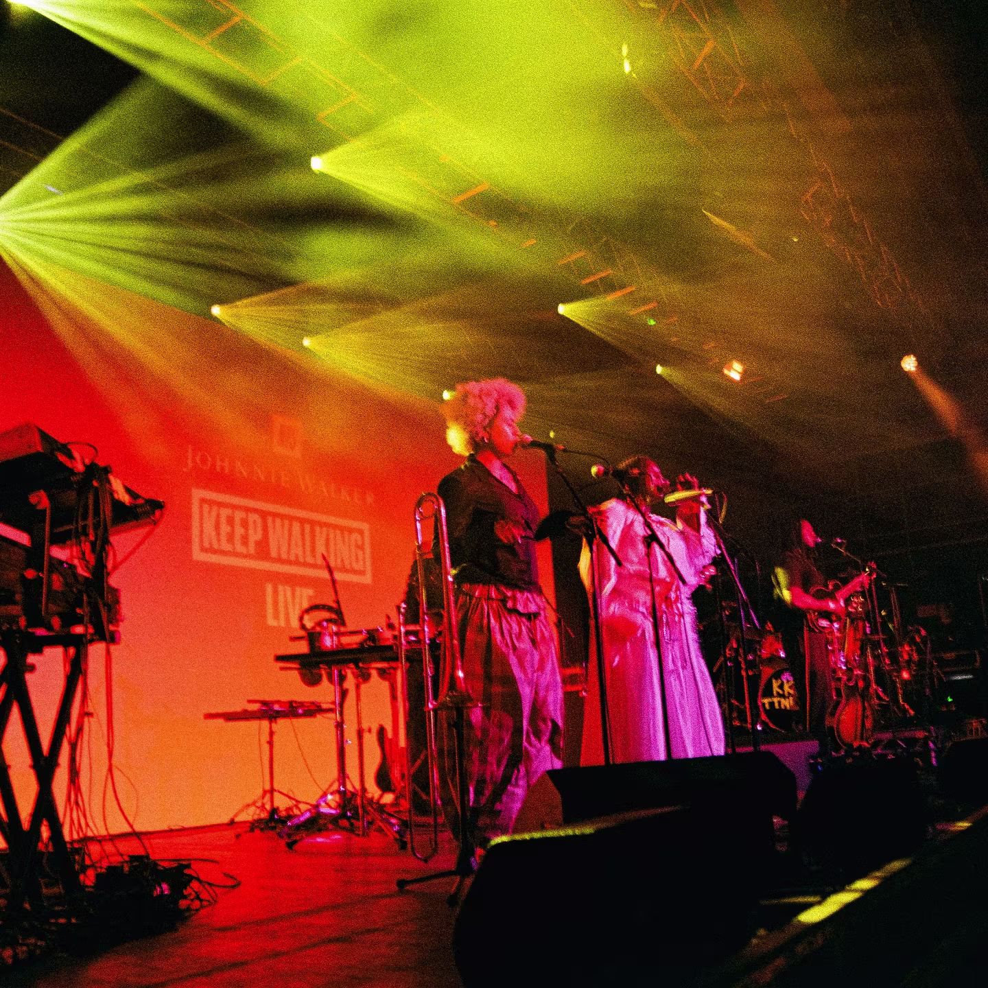 A band performs on stage with bright yellow lights and musical instruments, under a Keep Walking Live sign.