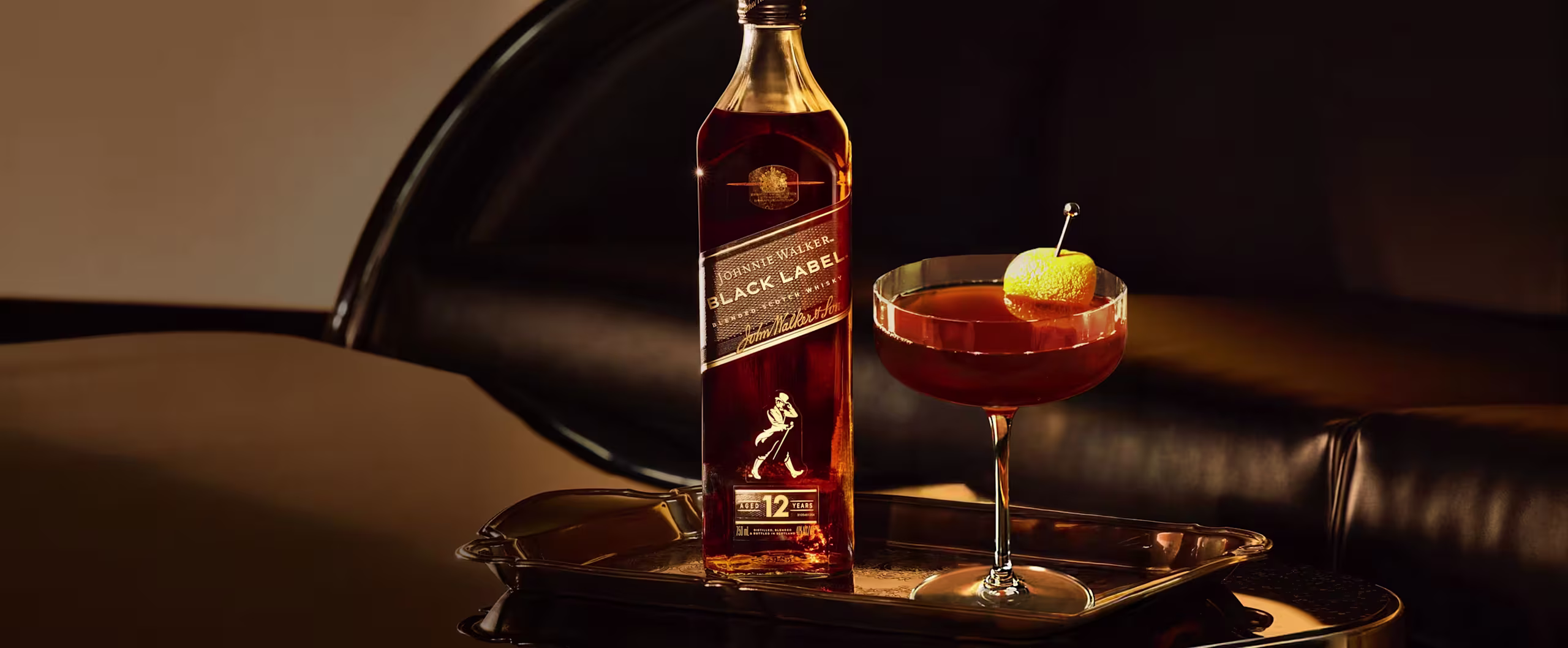 A bottle of Johnnie Walker Black Label whisky and a cocktail garnished with a lemon twist sit on a tray in a dimly lit, elegant setting with black leather furniture in the background.