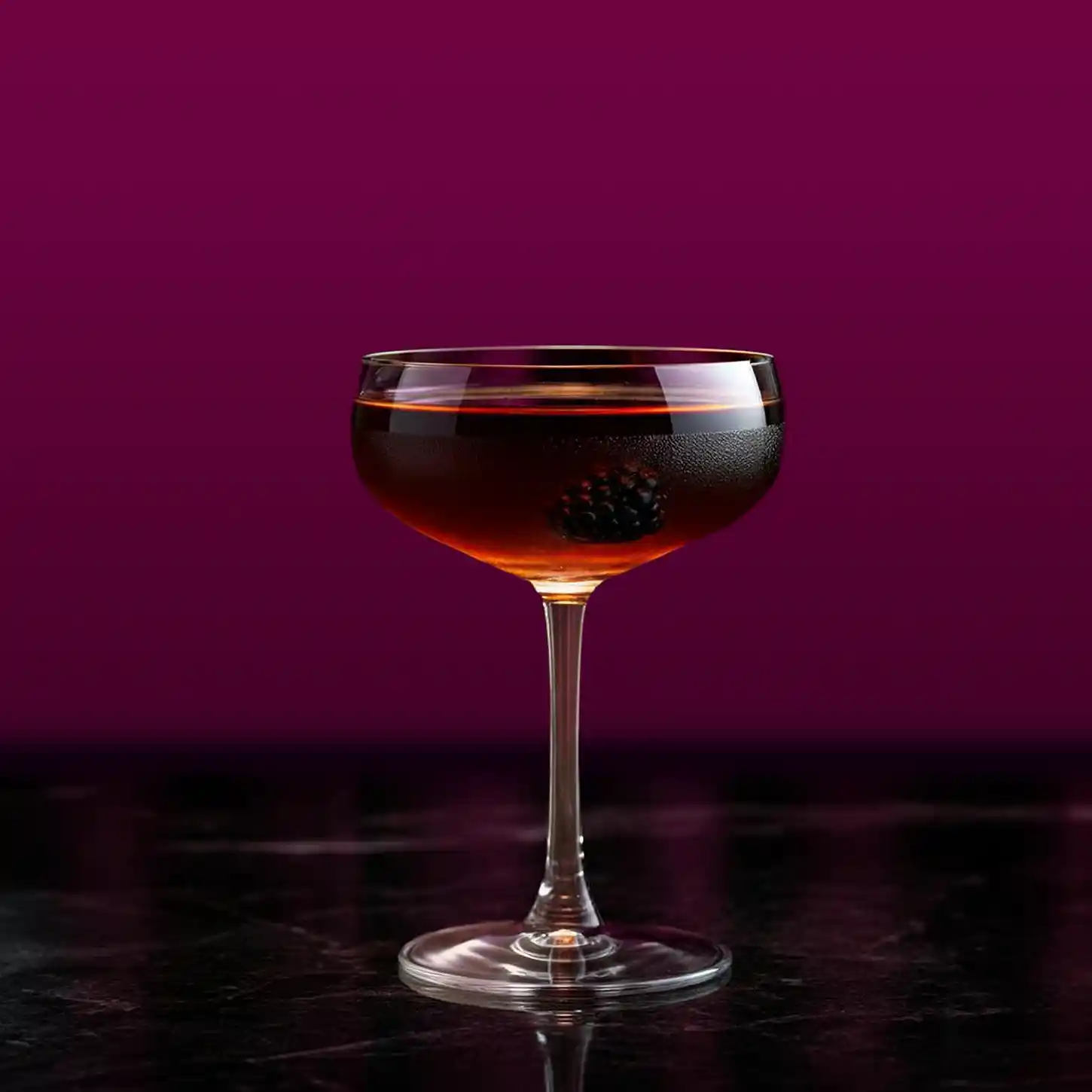 A dark cocktail garnished with a blackberry sits in a coupe glass against a deep purple background. The glass rests on a dark marble surface, creating a sophisticated and elegant presentation.