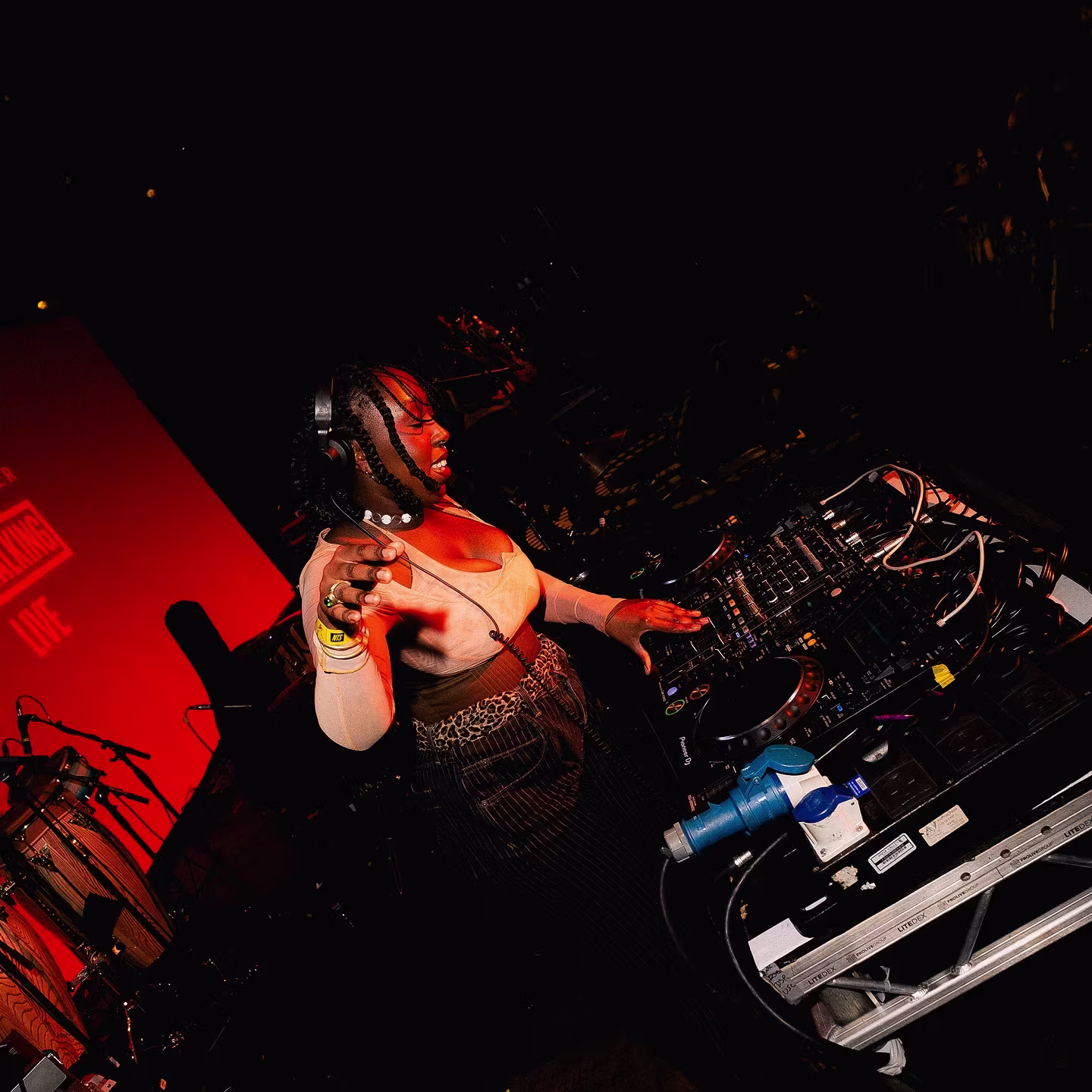 A DJ wearing headphones performs at a dark venue, illuminated by red stage lights.