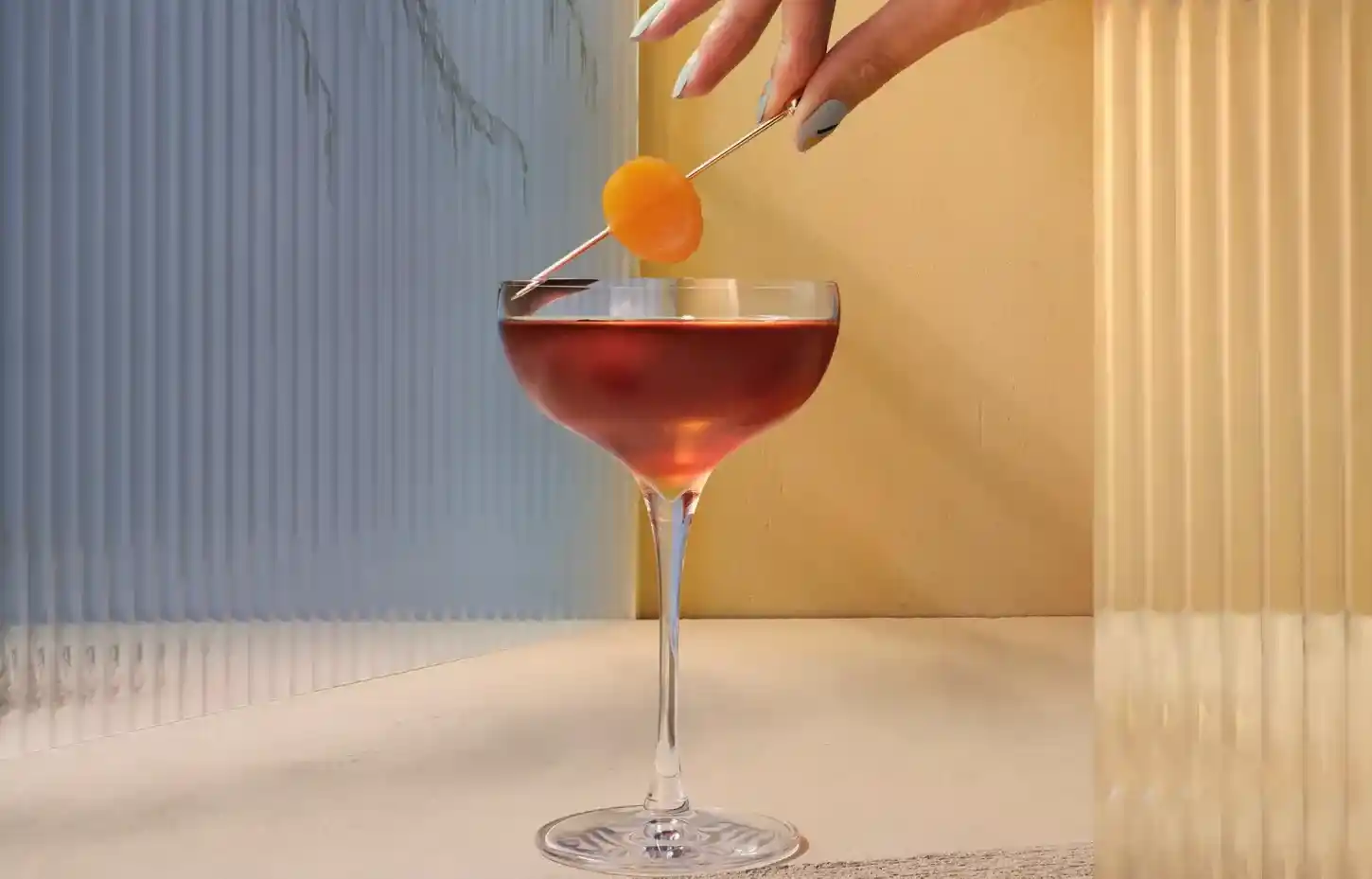 A hand is holding a toothpick with an orange fruit on it above a glass of red liquid. 
