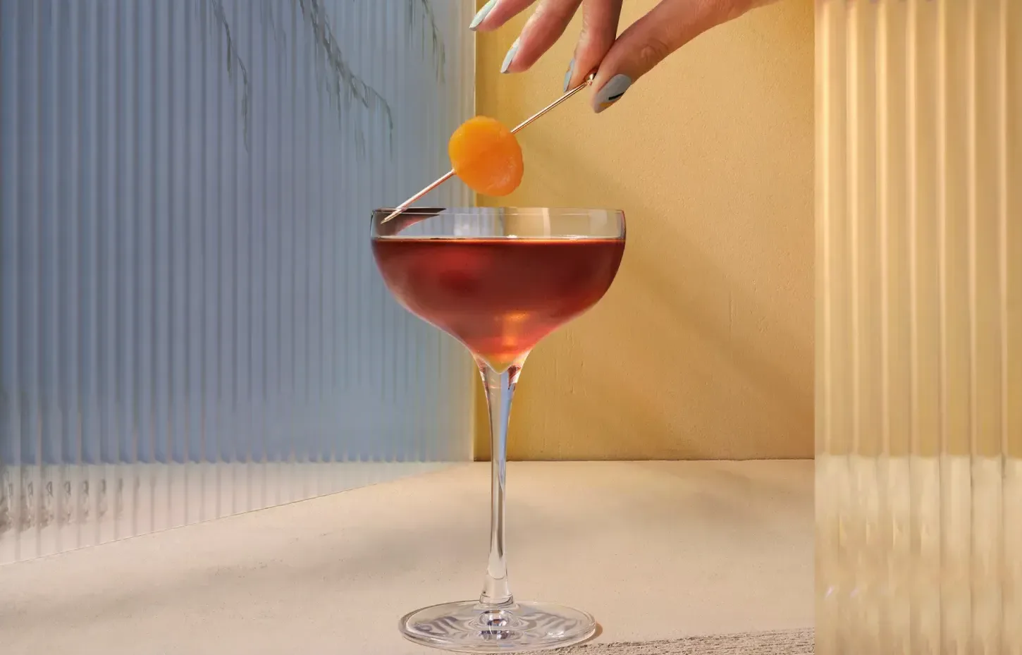 The Smooth & Floral Old Fashioned. A hand delicately holds a skewer with an orange garnish above a cocktail in a stemmed glass. The setting features textured glass panels casting shadows and a neutral, warm background.