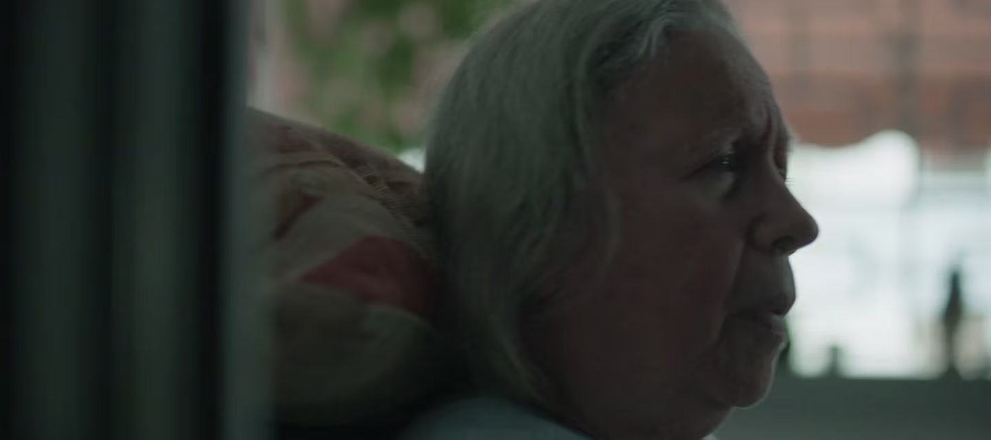 Se ve a una mujer mayor de cabello gris de perfil, pensativa, sentada en una habitación con una luz tenue. Tiene una almohada o cojín apoyado en su hombro. El fondo muestra una vegetación borrosa fuera de una ventana.