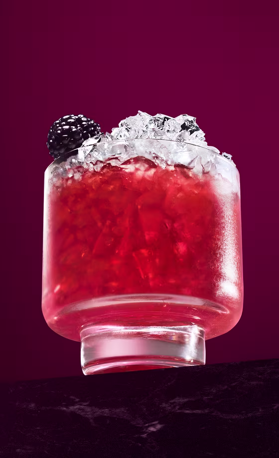 Un vaso corto y transparente lleno de hielo picado y un cóctel rojo vibrante, adornado con una mora encima, sobre un fondo magenta oscuro y colocado sobre una superficie oscura.