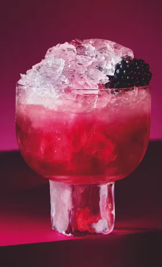 A glass of red cocktail filled with crushed ice and garnished with a blackberry, set against a deep red background with dramatic lighting.