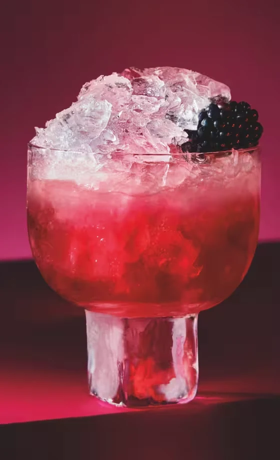 A glass of red cocktail filled with crushed ice and garnished with a blackberry, set against a deep red background with dramatic lighting.