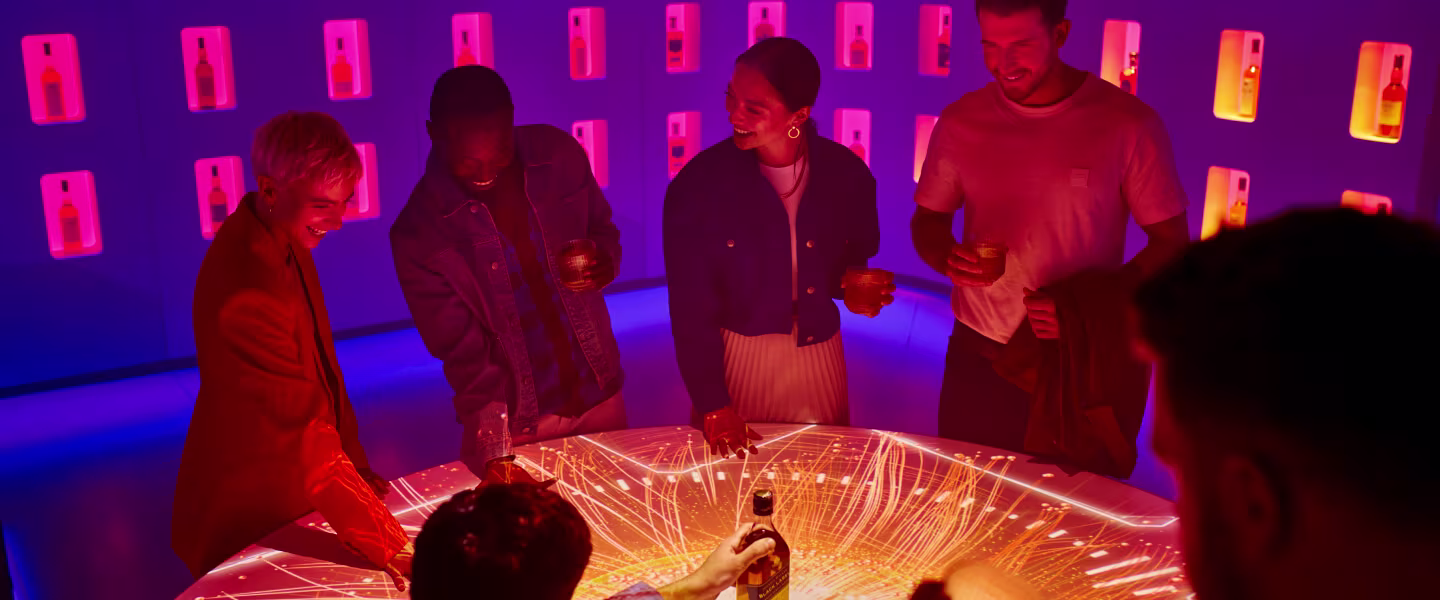 Un groupe de personnes se tient autour d'une table lumineuse avec un écran d'affichage numérique. Dans une pièce tamisée aux étagères éclairées, ils discutent autour d'un verre, créant une atmosphère animée et colorée.