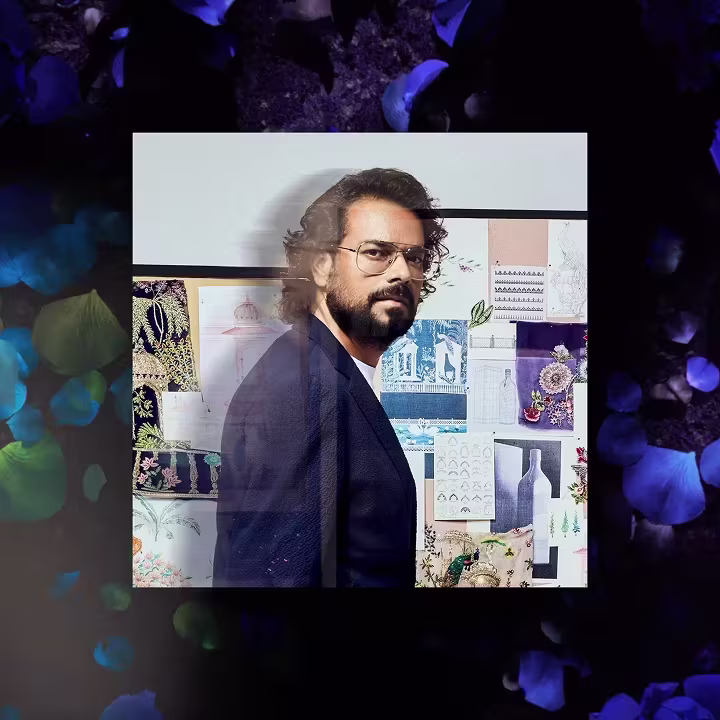 Rahul Mishra with glasses and a beard in front of a wall covered with sketches and fabric samples.