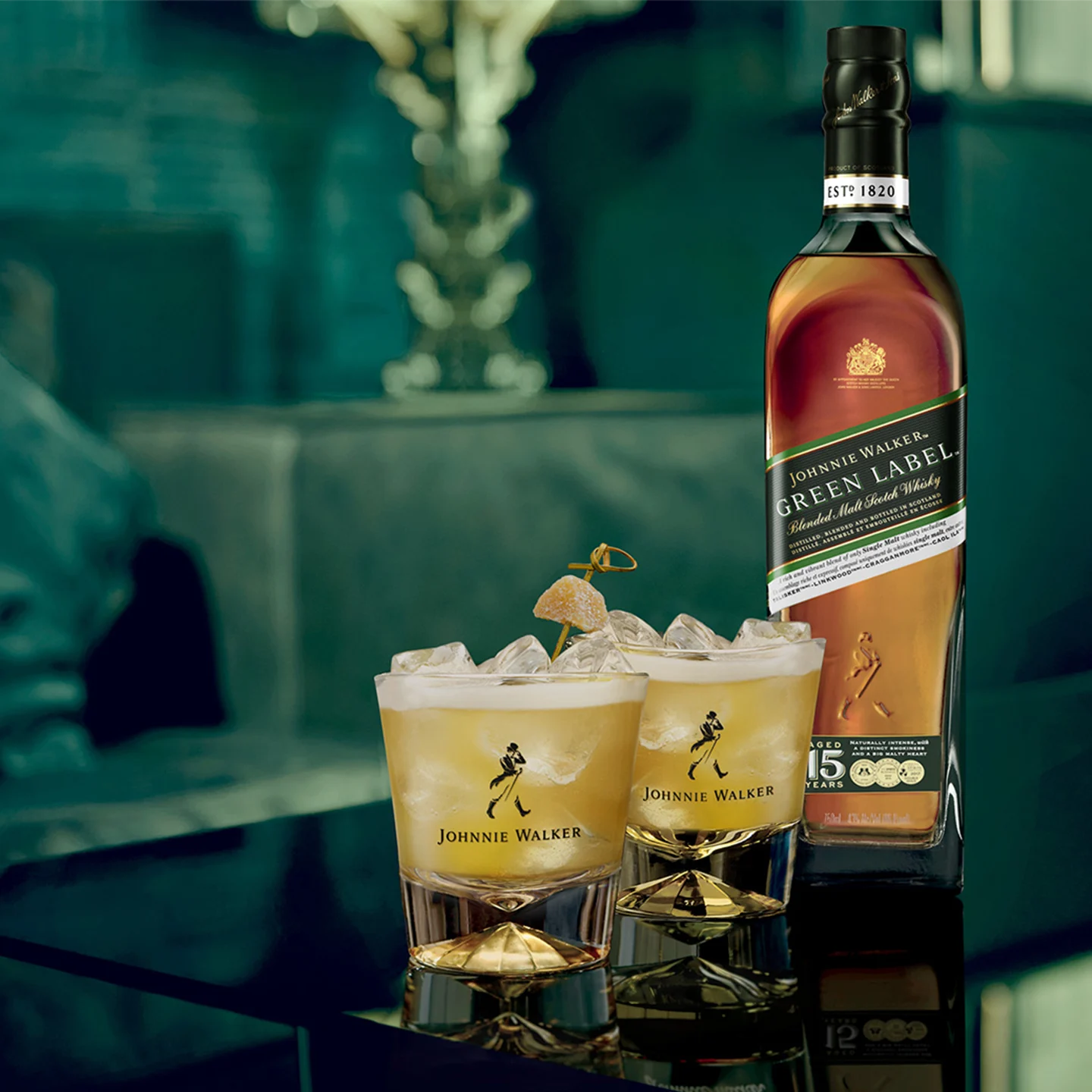 Ένα μπουκάλι σκωτσέζικο ουίσκι Johnnie Walker Green Label τοποθετείται σε ένα τραπέζι. Δύο ποτήρια γεμάτα με ουίσκι, πάγο και γαρνιτούρες αχλαδιών κάθονται κοντά. Το καπάκι του μπουκαλιού στηρίζεται στο τραπέζι και το φόντο είναι απαλά θολό.