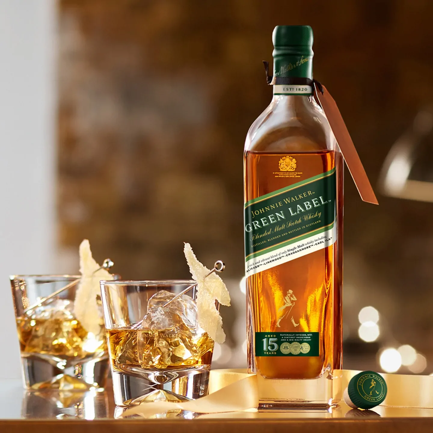 A bottle of Johnnie Walker Green Label whiskey stands on a table next to two glasses filled with whiskey and ice. A whiskey bottle cap lies nearby. The background is softly blurred, creating a warm, inviting atmosphere.