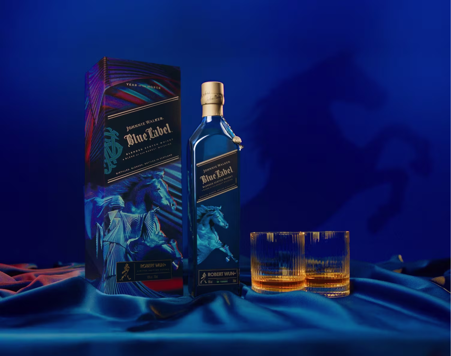 Blue Label whisky bottle, box, and two filled glasses on blue fabric with a horse shadow in the background.