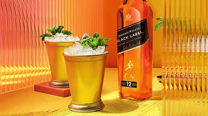 A hand holds a gold cup filled with crushed ice and mint beside a bottle of Johnnie Walker Black Label whisky on a vibrant yellow-orange background.