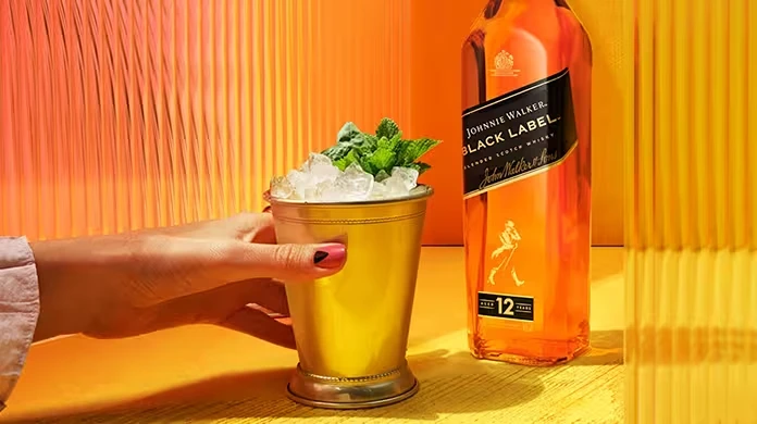 A hand holds a gold cup filled with crushed ice and mint beside a bottle of Johnnie Walker Black Label whisky on a vibrant yellow-orange background.