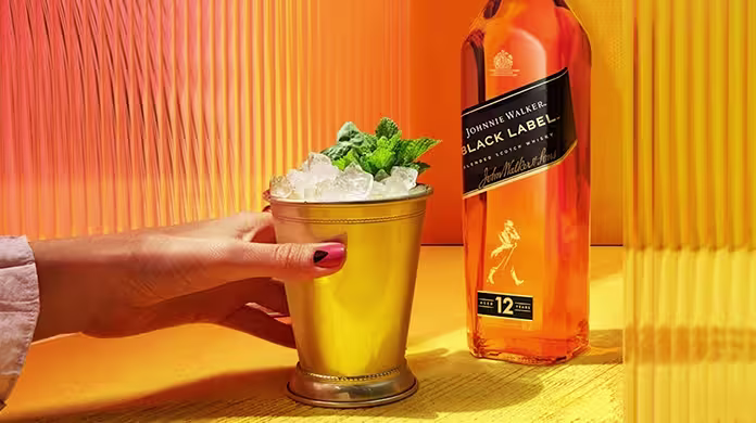  Ruka drží zlatý pohár naplněný drceným ledem a mátou vedle lahve whisky Johnnie Walker Black Label na zářivě žlutooranžovém pozadí.