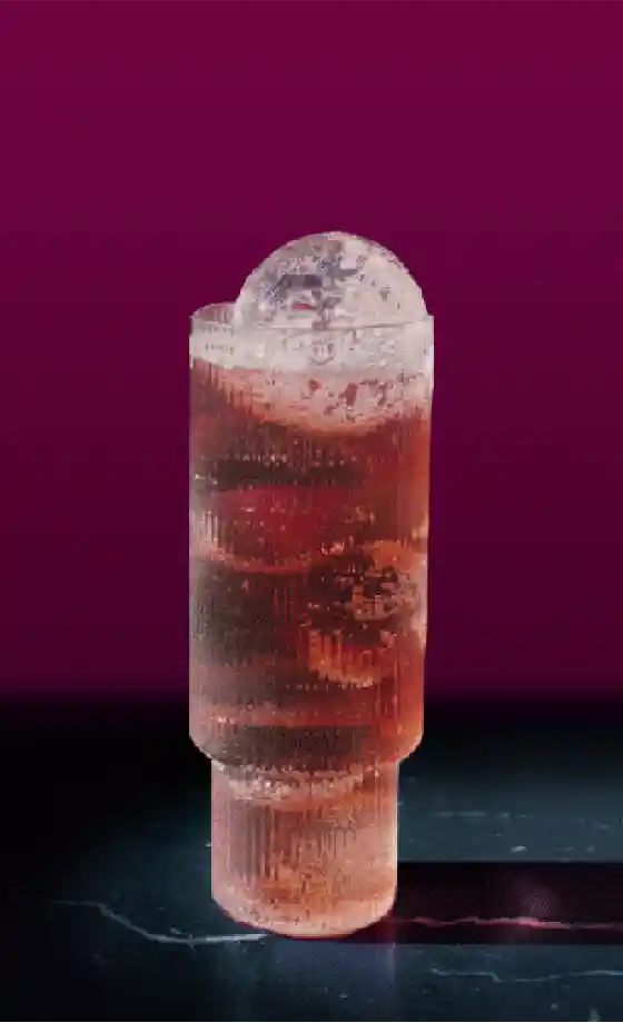 A tall, textured glass filled with a fizzy pink drink sits on a dark marble surface. The drink is topped with a large, clear spherical ice cube. The background is a deep burgundy color.