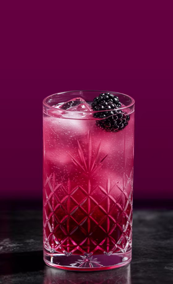 Un vaso alto grabado, lleno de una bebida gaseosa rosa y cubitos de hielo. Decorado con una mora fresca, reposa sobre una superficie oscura sobre un fondo morado intenso.